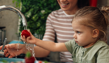 A mother smiling as her young daughter washes fresh strawberries in the sink.