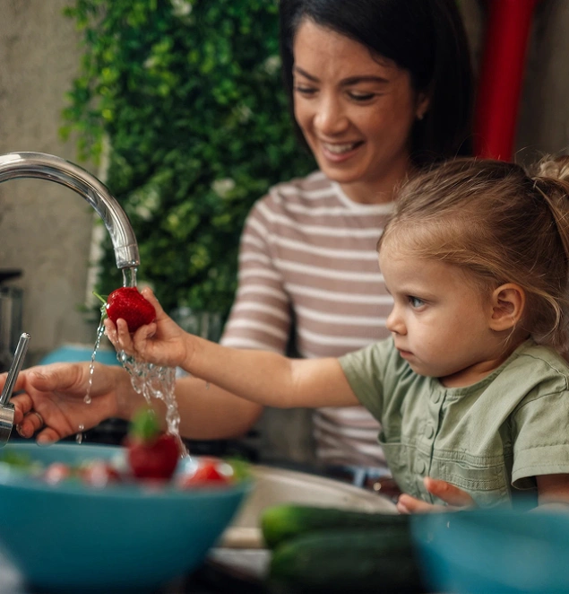 A mother smiling as her young daughter washes fresh strawberries in the sink.