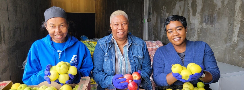 Three women smiling while holding fresh yellow and red apples over crates.