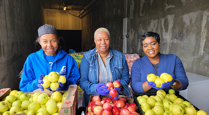 Three women smiling while holding fresh yellow and red apples over crates.